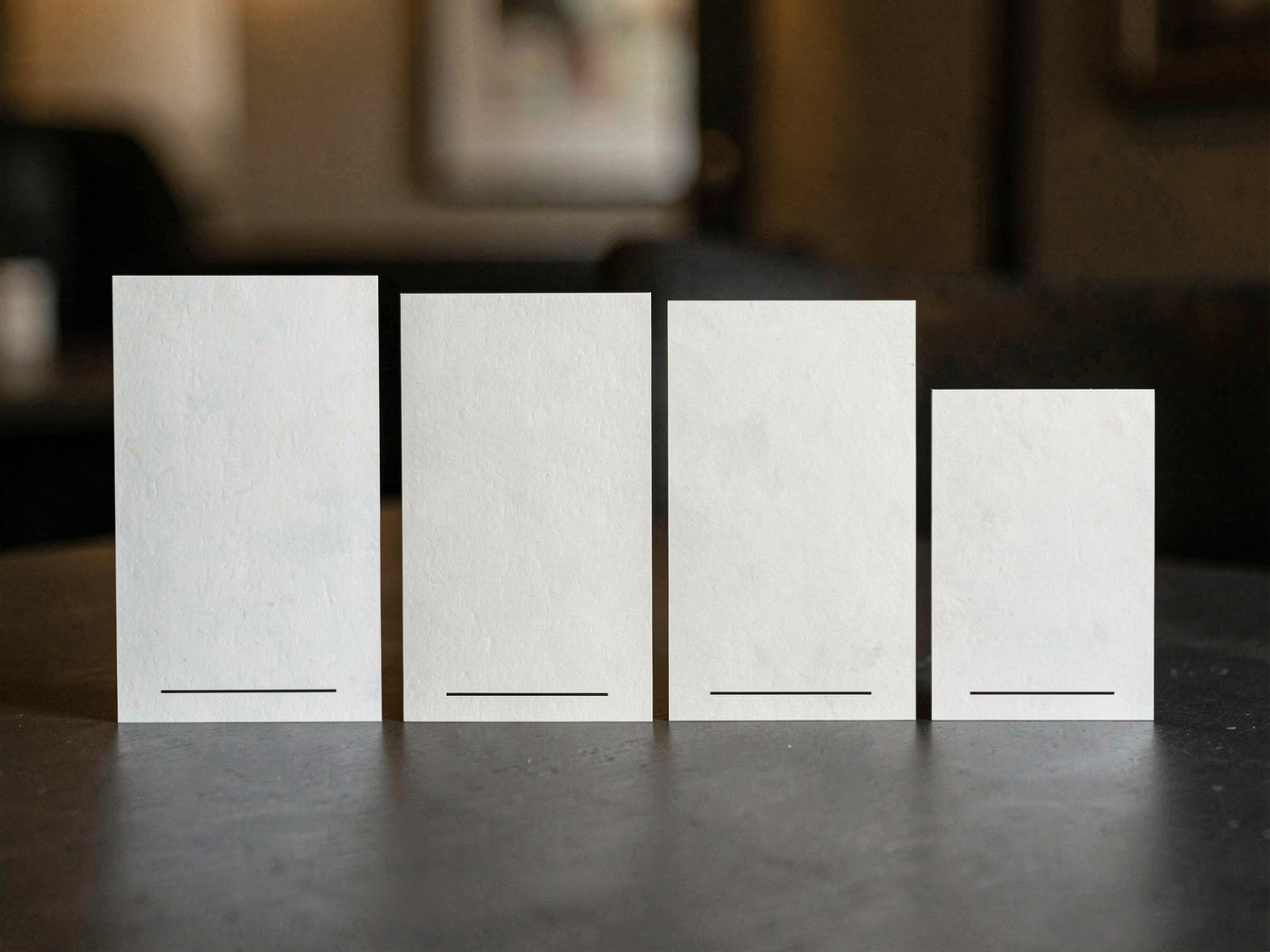 Vier leere Weinetiketten unterschiedlicher Größe nebeneinander auf einem dunklen Tisch, jedes mit einer schmalen leeren Zeile am unteren Rand